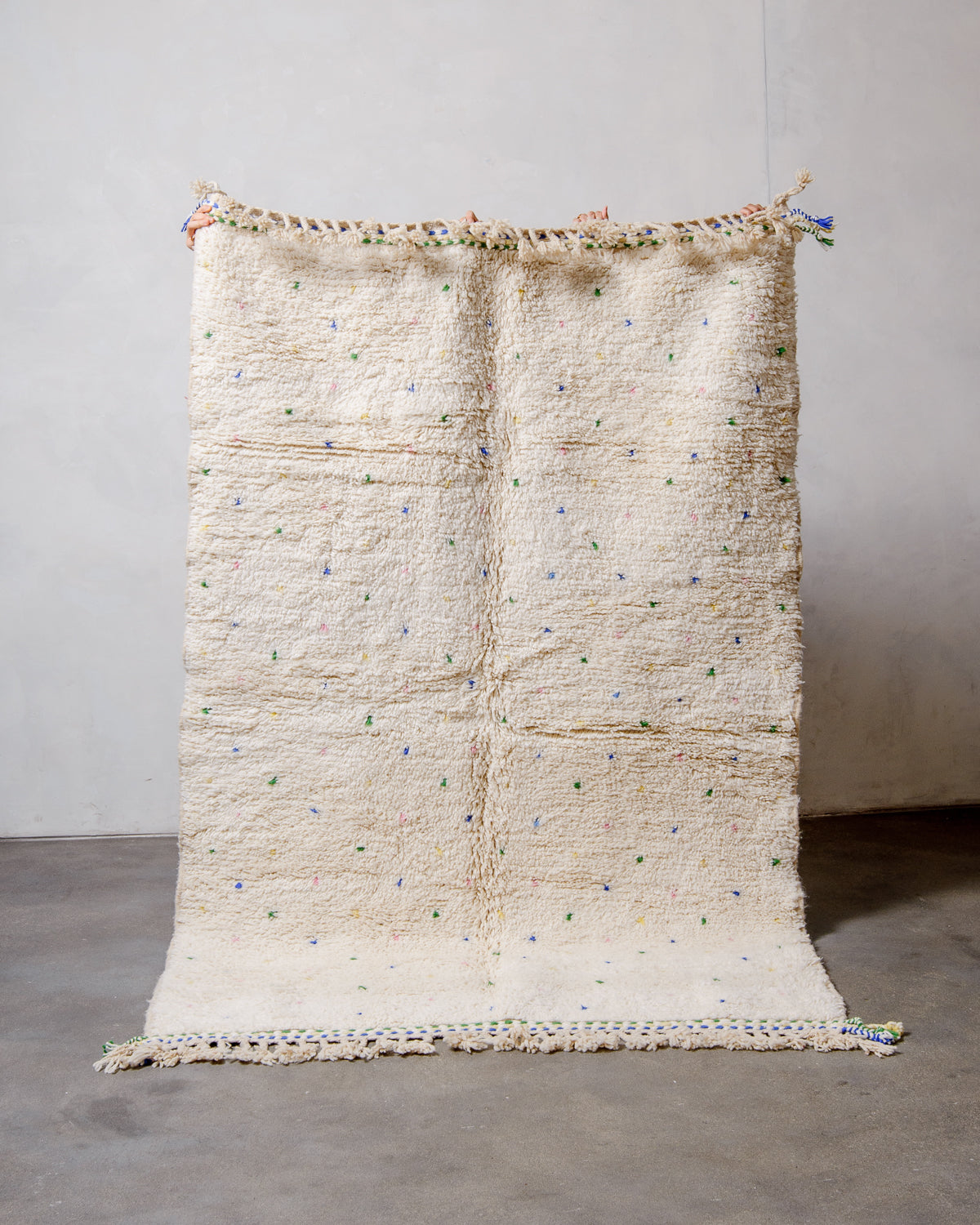 Berberlin Beniourain rug in cream wool with coloured dots being held up in front of a white wall.